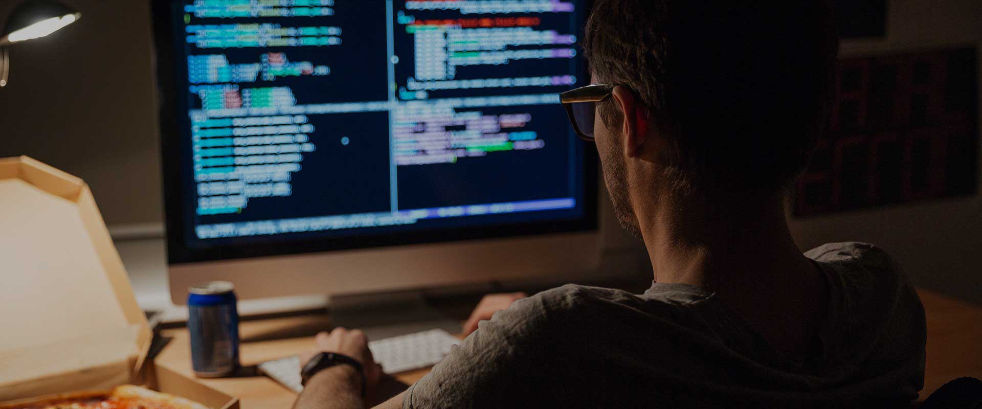 Back view of focused young programmer in glasses writing code and eating pizza